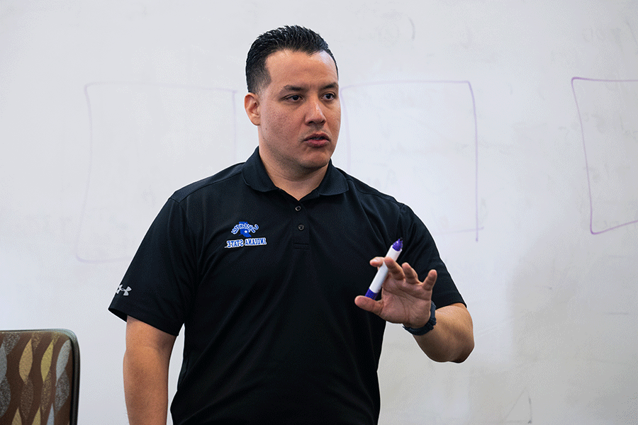 A male wearing a black polo holds a marker and stands in front of a white board while speaking