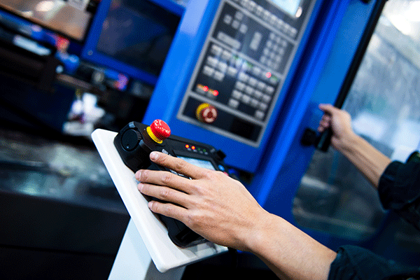 A pair of hands are shown engaging with a blue control panel. The right hand rests on a black control pad with two illuminated orange lights and a red button on a yellow base, and a few other buttons and lights on the far side. The pad sits on a white pedestal. In the background, a tall blue control panel with various buttons, lights, and a red knob is also visible. The left hand in the image rests further away on a black vertical handle attached to the control panel.