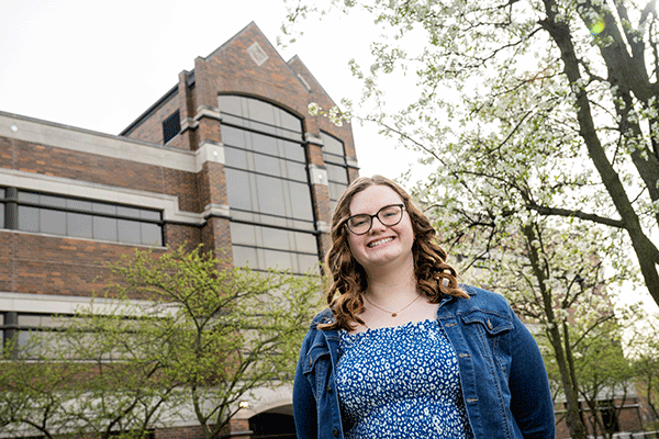 A photo of a white female student with brown hair and glasses, who stands at a distance in front of a tall brick building with many glass windows. It is the John T. Myers Technology Center. The woman is wearing a blue dress with a white pattern on it and a blue jean jacket. Trees with white flowers and dark branches are visible in the background.