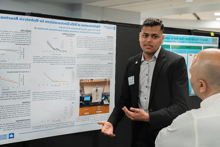 A male student of color with short black hair, wearing a black suit jacket and gray, open-collared shirt, presents his Honors research poster to an onlooker, an older man with a bald head and wearing a white shirt. The poster depicts photos, charts, and graphs related to the student’s research. Another blue, green, and white poster is visible in the background.   