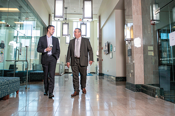 A WHITE PROFESSOR AND WHITE STUDENT, BOTH IN PROFESSIONAL ATTIRE, WALKING TOWARDS THE CAMERA AND TALKING IN A BRIGHTLY LIT HALLWAY. 