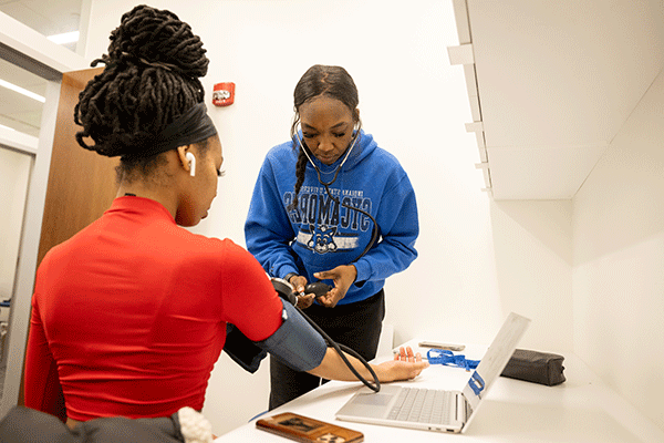 Two Black women pictured in a white exam room, with one measuring the other’s blood pressure. The woman taking the blood pressure wears black pants and a blue Sycamores sweatshirt with Sycamore Sam on the front. She has a stethoscope in her ears and is holding the pump to a blood pressure cuff in her hands. The woman wearing the blood pressure cuff is seated with her back to the camera. She wears a red top, a black bandanna, has a white earpiece in her ear, and her dreadlocked hair is piled atop her head.